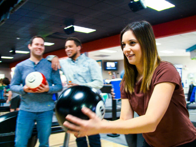 Young woman bowling a ball with two friends standing in the background watching and smiling