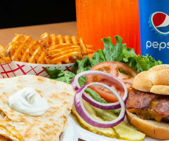 A plate of crisscut fries, a quesadilla, and a hamburger next to sodas